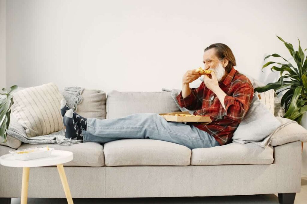 Older man in a red plaid shirt and jeans sits on a beige couch, eating pizza from a box with his feet up on the couch.