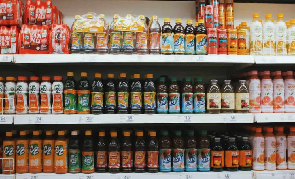Rows of bottled drinks on supermarket shelves, including fruit juices, iced tea, and energy drinks, with price tags displayed below each item.