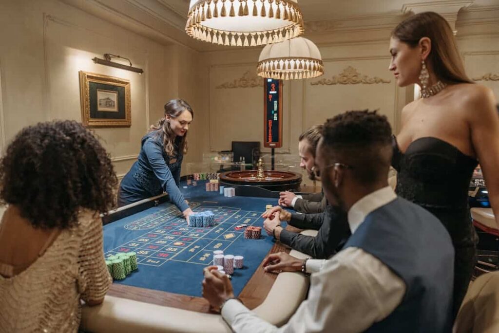 People are seated at a casino table, engaging in a game of roulette. A dealer manages the chips while the participants, dressed formally, watch and participate.