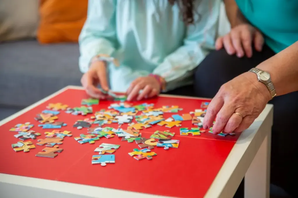 Two people, one adult and one child, work together on assembling a colorful jigsaw puzzle on a red tabletop.