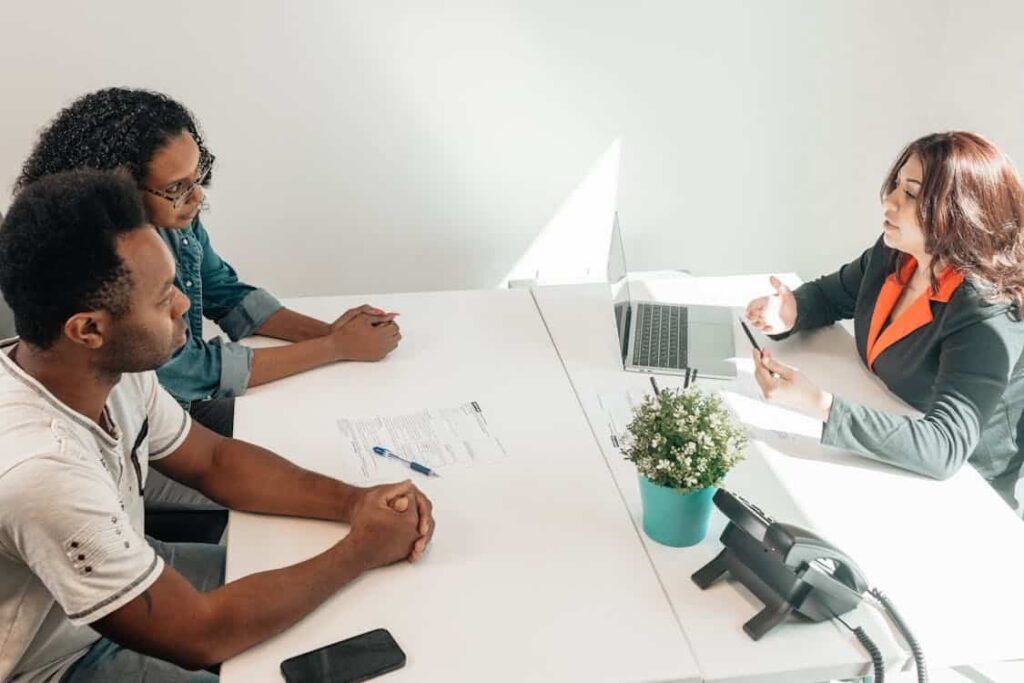 Three people having a discussion in an office setting, with paperwork, a laptop, and a potted plant on a white table.