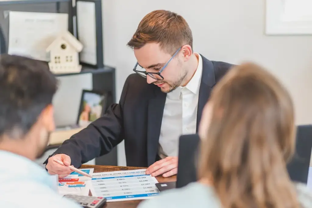 A man in a suit and glasses sits at a desk, pointing to a document with charts. Two other people are in the foreground, partially visible, engaged in discussion.