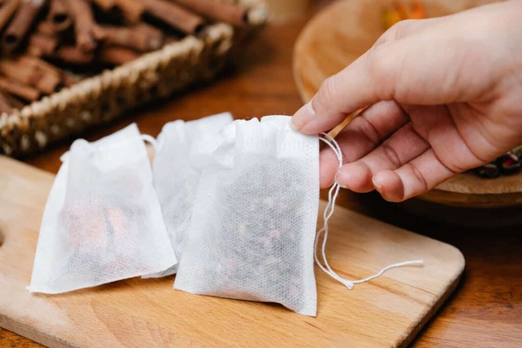 A hand holding a small white mesh bag containing dried herbs or tea leaves, with additional similar bags resting on a wooden surface.