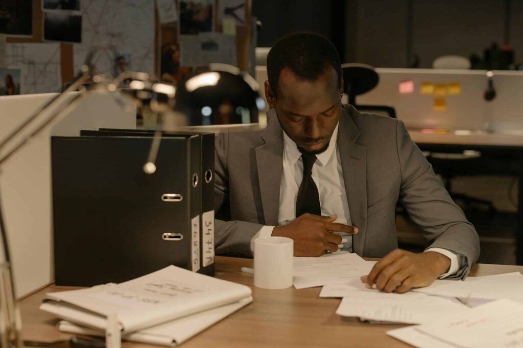 A man in a suit sits at a desk in a dimly lit office, reviewing documents. A white mug, ring binders, and papers are on the desk. In the background, a corkboard with notes and photos is visible.
