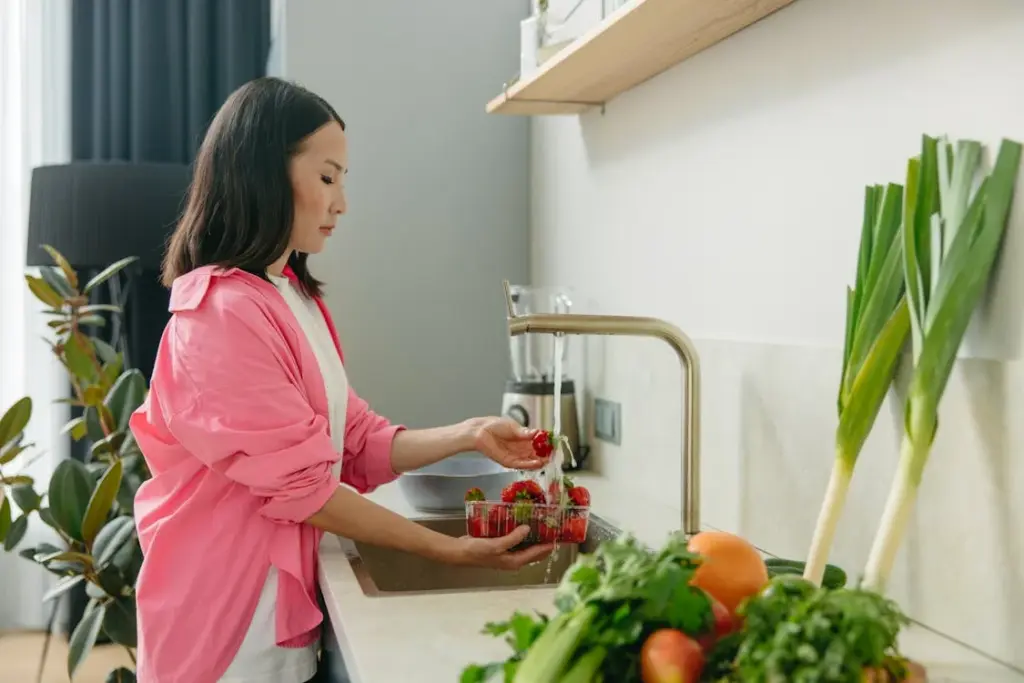 A woman in a pink shirt washes strawberries at a kitchen sink. Fresh vegetables are on the counter beside her.