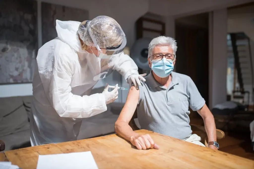 A healthcare worker in protective gear administers a vaccine injection to an older man seated at a table. Both are wearing face masks.