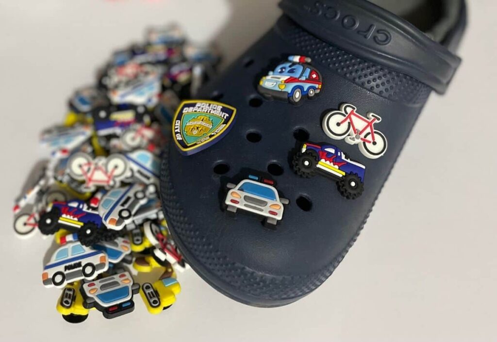 A navy blue Croc with various police-themed charms, including a police car, bicycle, shield, and truck, resting against a pile of similar charms on a white surface.
