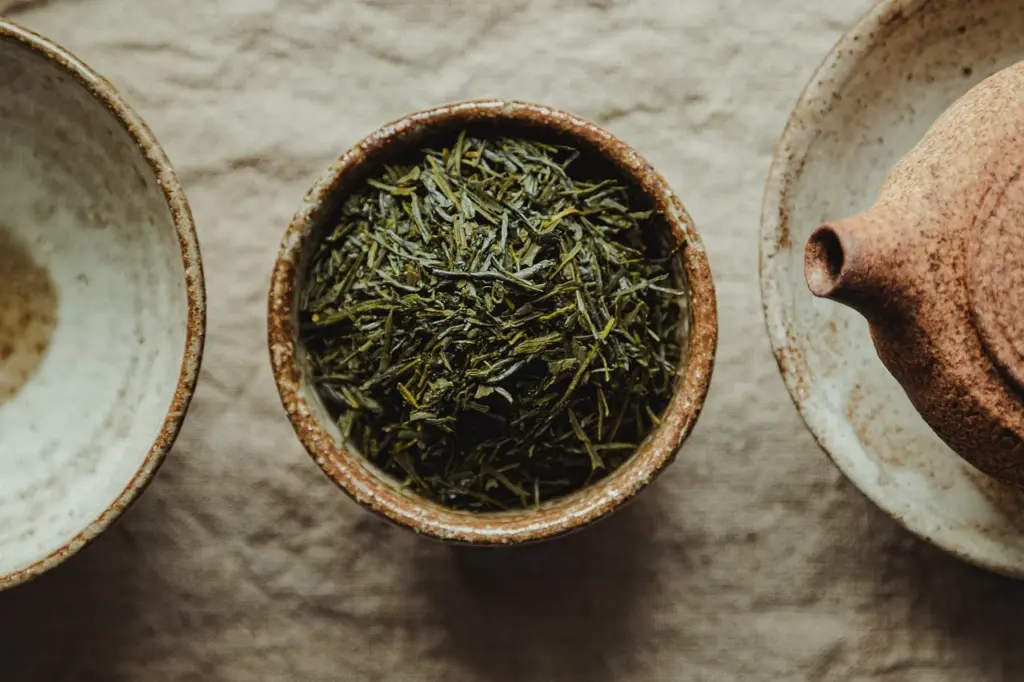 A bowl filled with loose green tea leaves placed on a beige cloth, flanked by a saucer on the left and a clay teapot on the right.