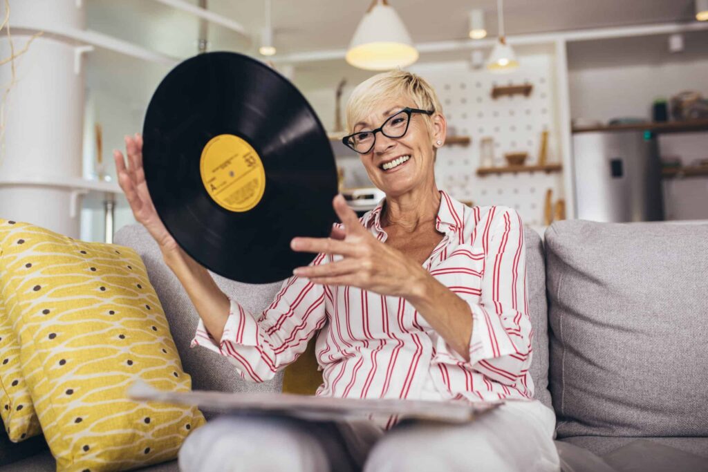 A woman with glasses and short blond hair, sitting on a couch, is holding up a vinyl record and smiling.