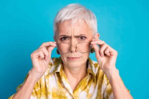 Older person with short gray hair and a serious expression adjusts clear glasses with both hands, wearing a yellow patterned shirt against a bright blue background.
