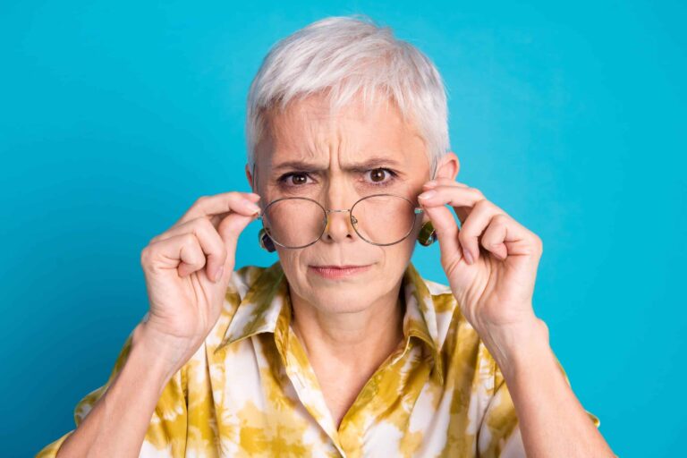 Older person with short gray hair and a serious expression adjusts clear glasses with both hands, wearing a yellow patterned shirt against a bright blue background.