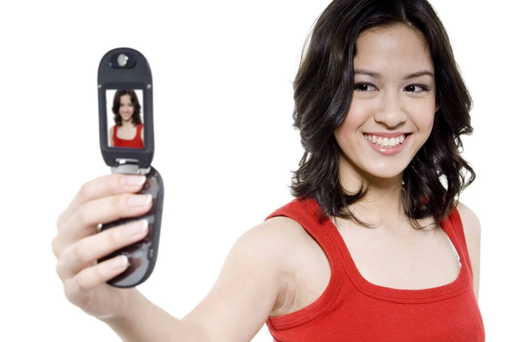 A smiling woman in a red tank top takes a selfie using a flip phone against a plain white background.