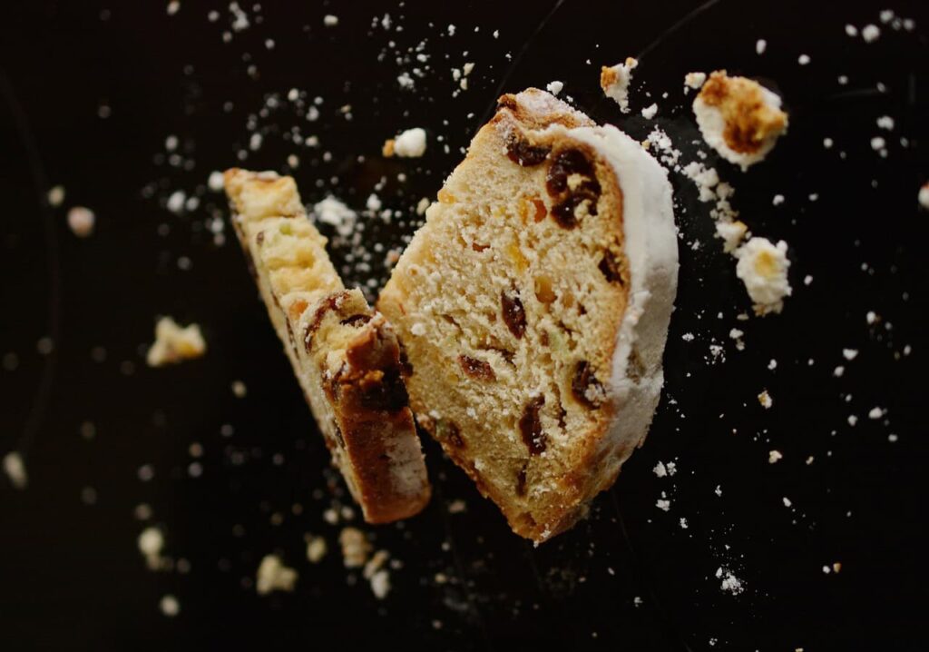 Two slices of fruit and nut bread with powdered sugar crust on a dark surface with crumbs scattered around.