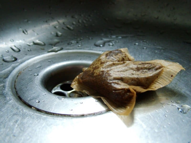 A used tea bag rests on the metal surface of a wet kitchen sink near the drain.