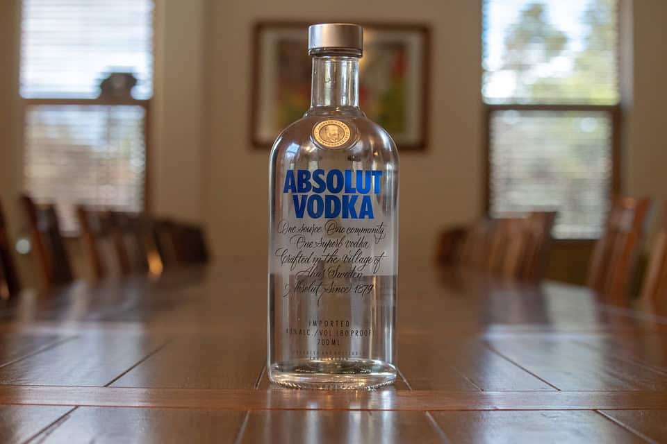 A bottle of Absolut Vodka sits on a wooden table in a room with large windows and wooden chairs in the background.