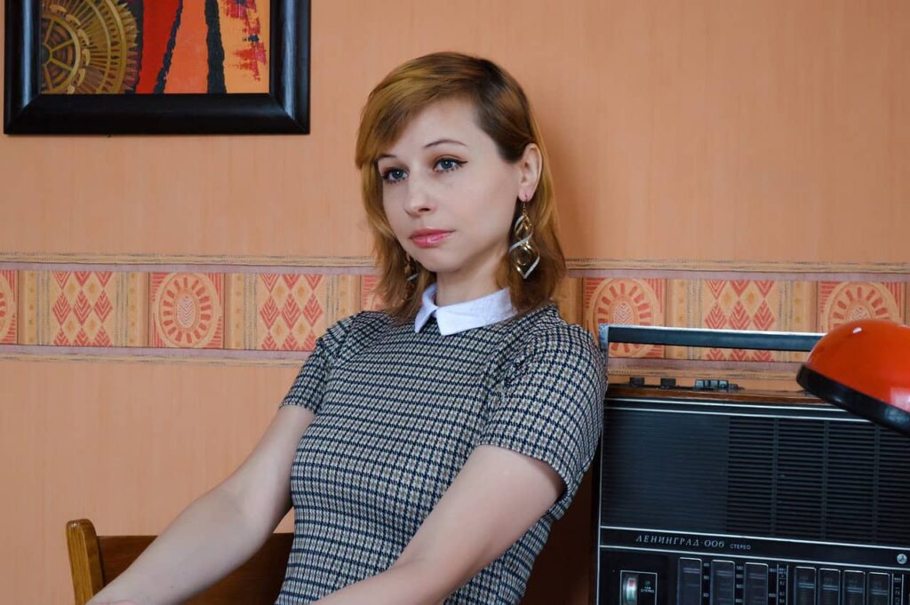 A woman with short, light brown hair wearing a checkered dress sits next to a vintage radio, with an orange wall and patterned wallpaper in the background.