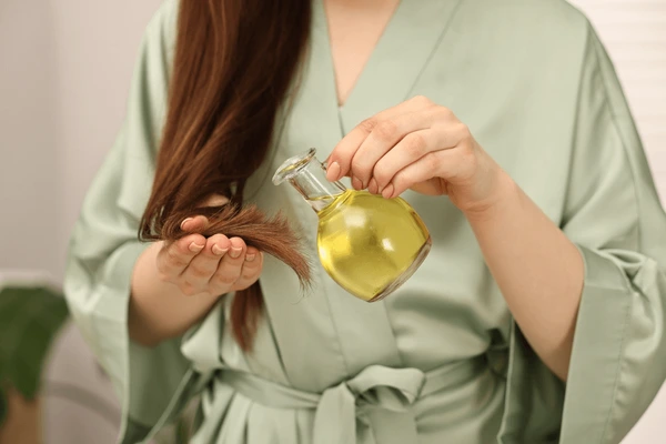 A person in a green robe applies oil to the ends of their long hair using a small glass bottle.