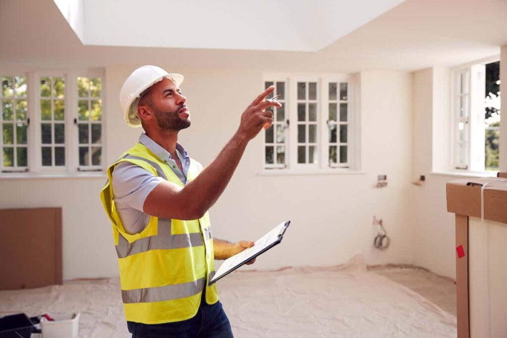 A construction worker in a yellow safety vest and hard hat holds a clipboard and points upward inside a partially renovated room.