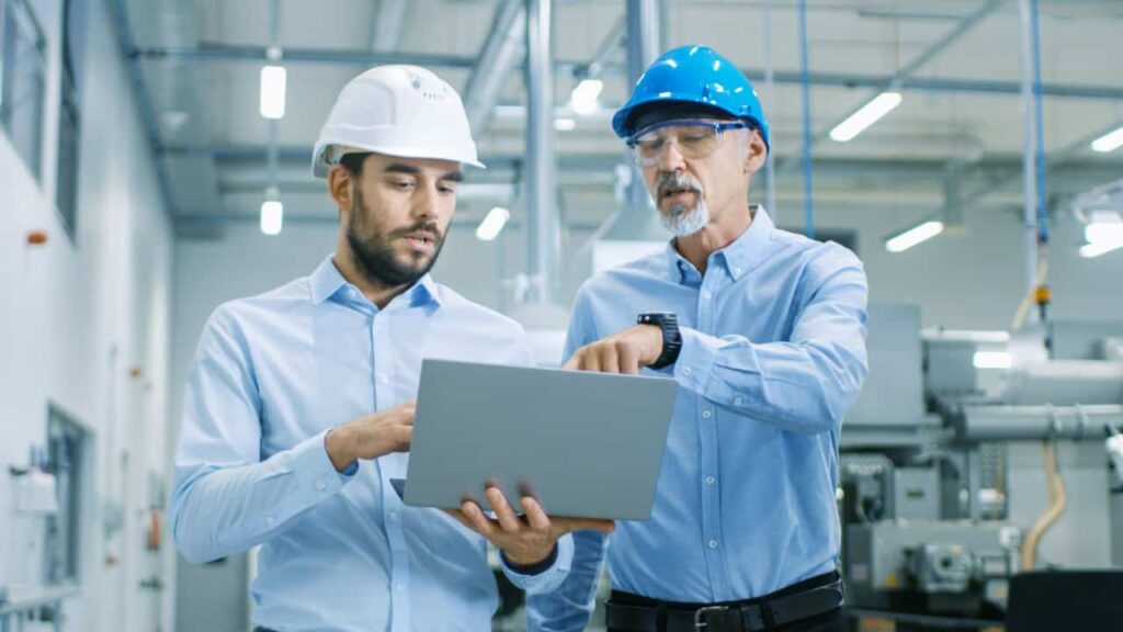 Two men in safety helmets stand in an industrial setting examining a laptop.