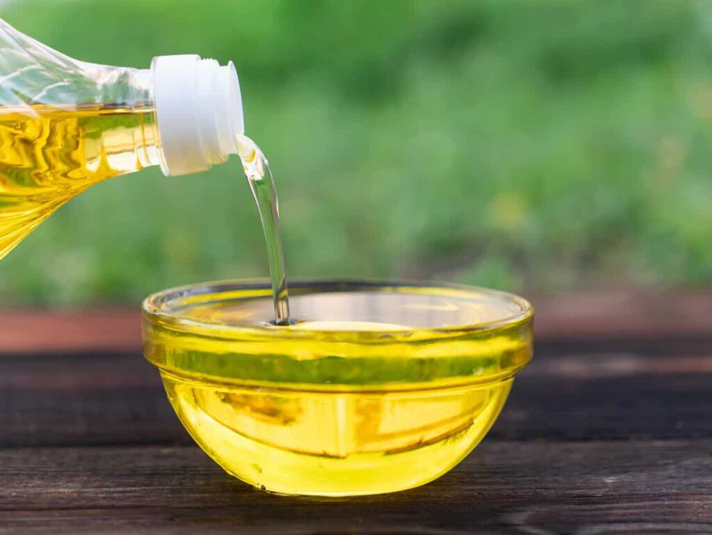 Clear liquid being poured from a bottle into a small transparent bowl on a wooden surface, with a blurred green background.