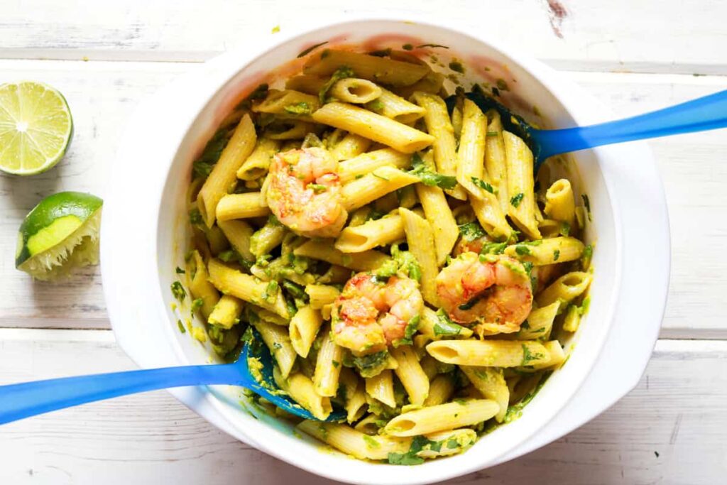 A bowl of penne pasta with shrimp and herbs, accompanied by slices of lime, with blue utensils.