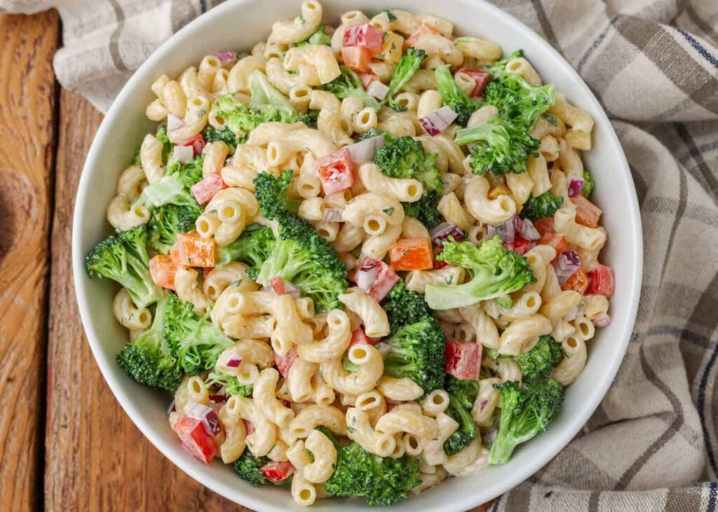 A bowl of macaroni salad with broccoli, red bell peppers, and red onions, dressed in a creamy sauce.