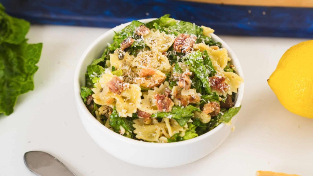 A bowl of pasta salad with bow-tie pasta, lettuce, bacon pieces, and grated cheese served on a table next to a lemon and a spoon.