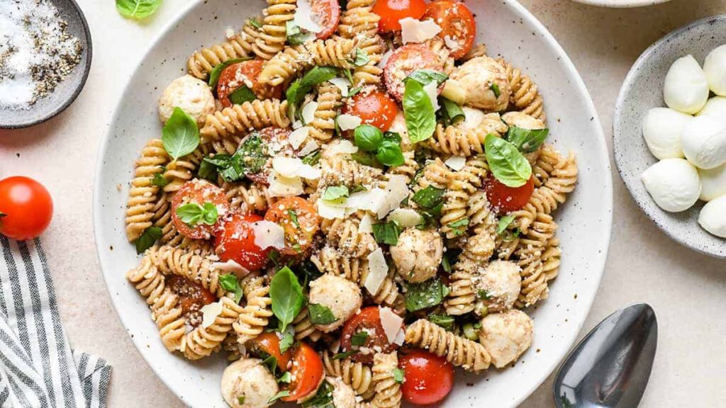 Plate of fusilli pasta salad with cherry tomatoes, mozzarella balls, spinach, chicken meatballs, and basil, garnished with cheese shavings.