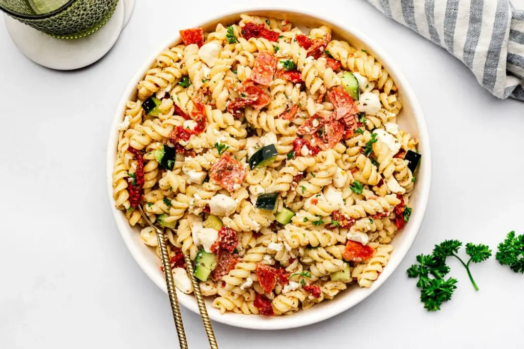 A bowl of pasta salad with fusilli, chopped vegetables, feta cheese, and herbs on a white surface, accompanied by a fork and napkin.
