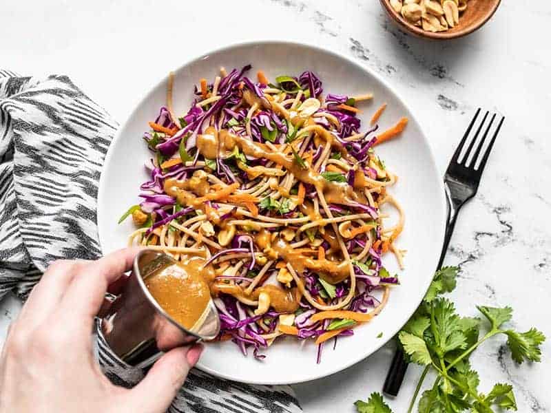 A hand pouring peanut sauce over a salad with purple cabbage, carrots, and bean sprouts on a white plate, accompanied by a fork, a bowl of peanuts, and cilantro.