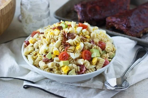 A bowl of pasta salad with rotini, cherry tomatoes, diced avocado, boiled eggs, bacon, and dressing. A tray with grilled meat is in the background. A fork and spoon rest nearby.