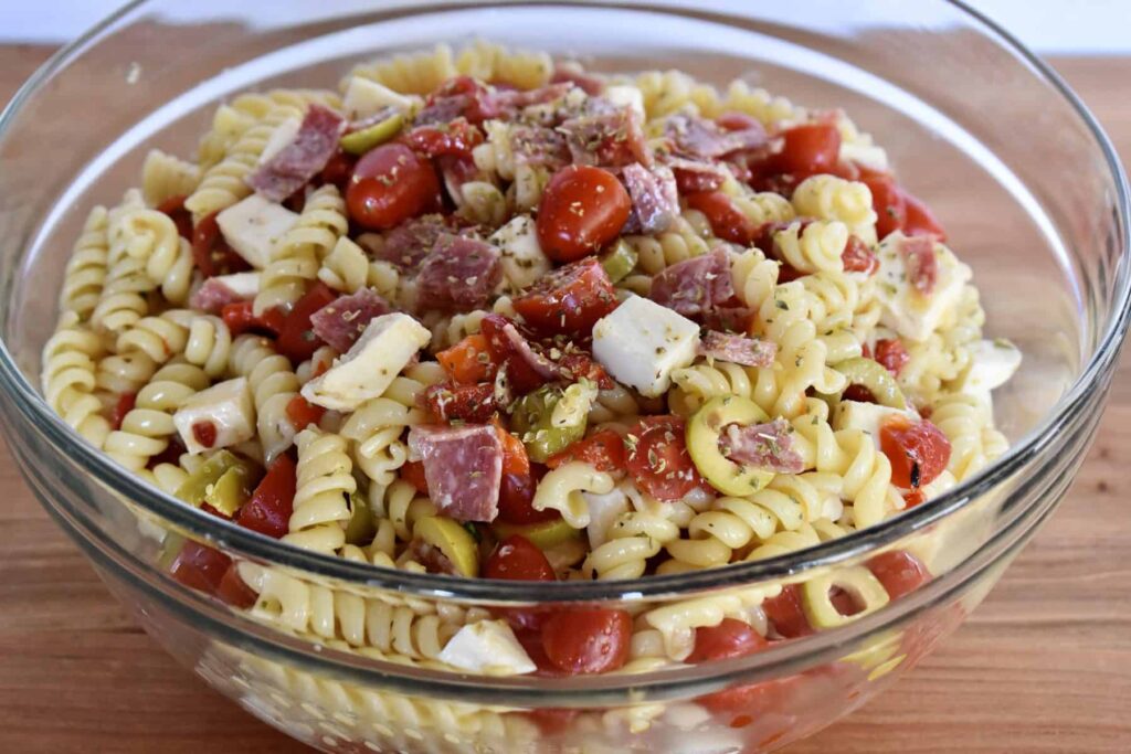 A glass bowl filled with rotini pasta salad, featuring cherry tomatoes, cheese cubes, salami pieces, and herbs.