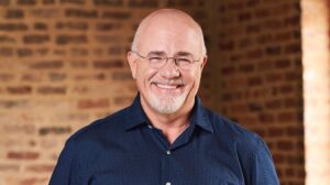 Smiling bald man with glasses and a goatee, wearing a blue shirt, standing in front of a brick wall.