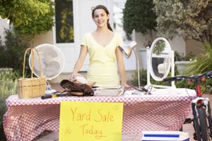 Person standing behind a table with various items and a "Yard Sale Today" sign on it.