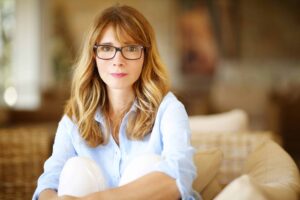 A woman with long hair and glasses sits on a couch, wearing a light blue shirt and white pants, looking directly at the camera.