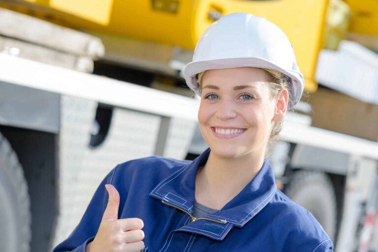 Woman in a hard hat gives a thumbs up while standing in front of industrial equipment.