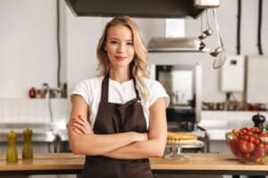 A woman in an apron stands confidently in a kitchen with arms crossed, with a pie and a bowl of tomatoes on the counter.
