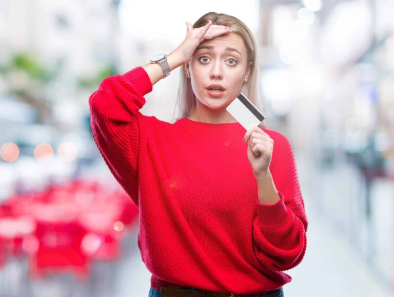 Woman in a red sweater holds a credit card, with one hand on her forehead, and an expression of surprise, standing in an outdoor setting with blurred background.