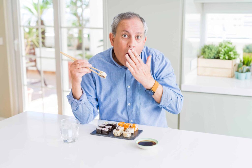 A man in a blue shirt sits at a table with sushi, holding chopsticks and covering his mouth in surprise. A glass of water and a dish of soy sauce are also on the table.