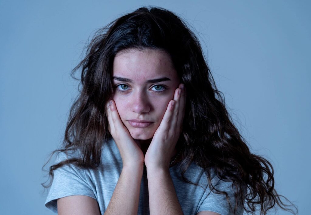 A person with long, wavy hair rests their face in their hands, looking directly at the camera with a neutral expression. They are wearing a gray shirt against a plain blue background.