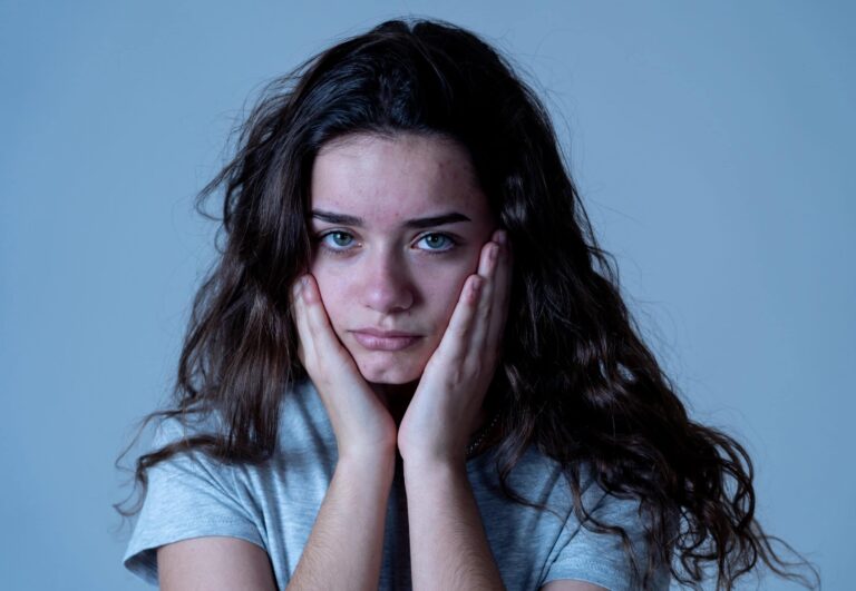 A person with long, wavy hair rests their face in their hands, looking directly at the camera with a neutral expression. They are wearing a gray shirt against a plain blue background.