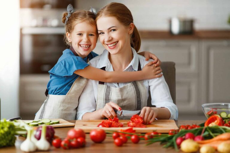 A woman and a child, both in aprons, smile while preparing vegetables in a kitchen.