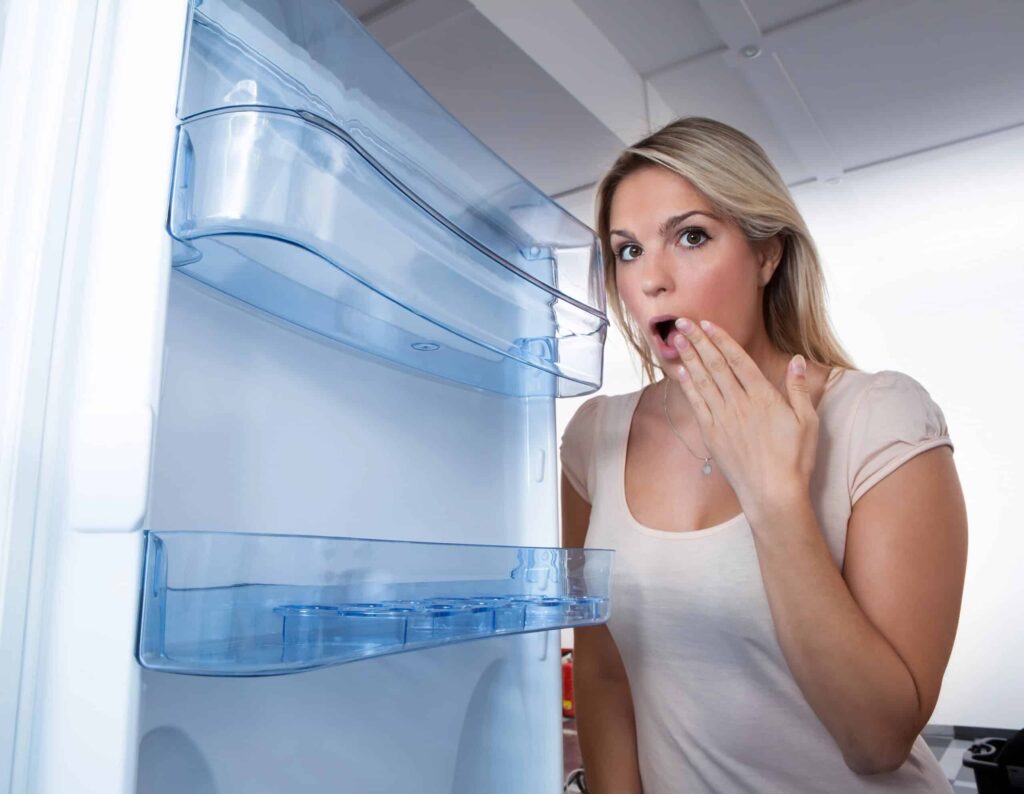 A surprised woman looks inside an open, empty refrigerator.