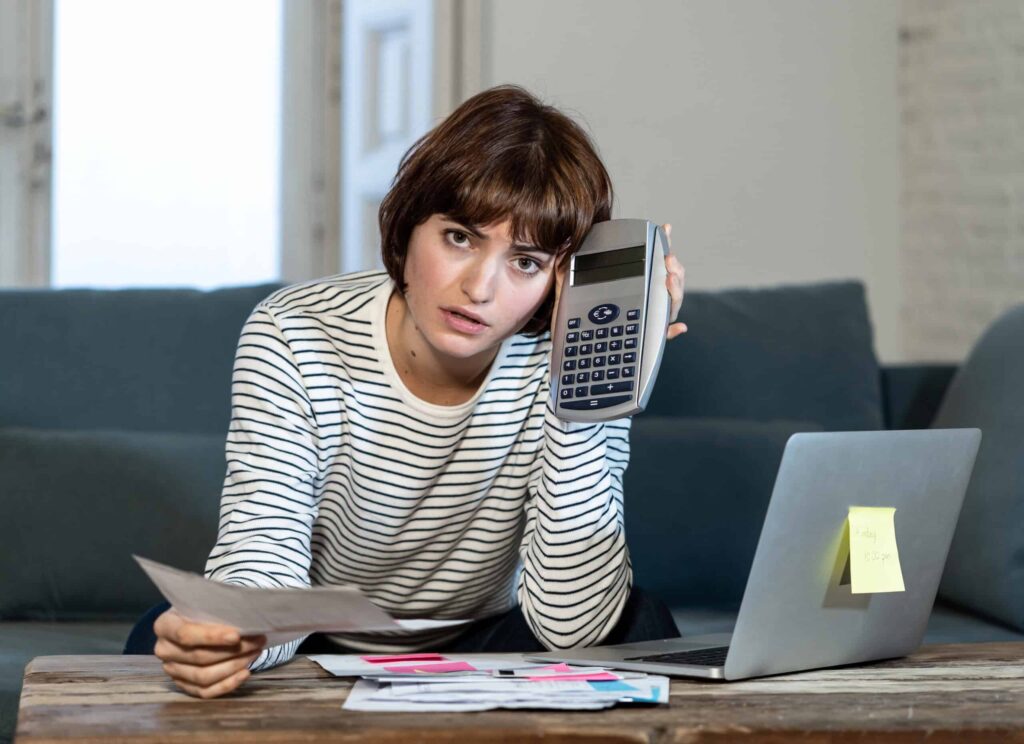 A person sits on a couch, looking serious, holding a calculator to their ear, with a laptop and papers on a table in front of them.