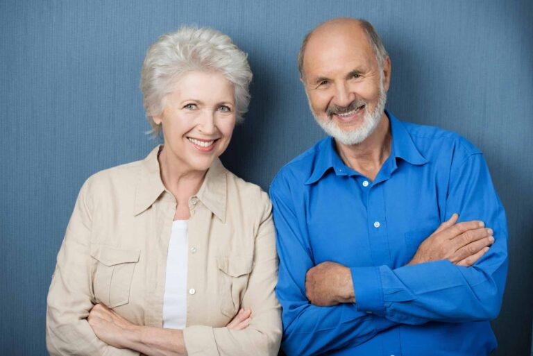 Older woman and man smiling, standing with arms crossed in front of a blue background.