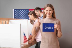 A woman smiling and holding an "I Voted" sign, with two people voting at booths in the background and a U.S. flag on the wall.