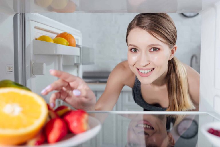A woman smiles while reaching into a refrigerator with citrus fruits on a plate in the foreground.