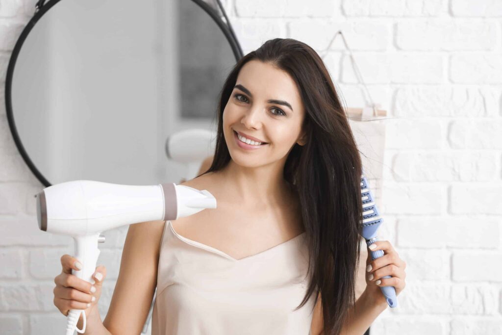 A woman is smiling while holding a hairdryer and a brush in front of a round mirror.