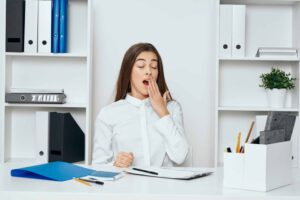 Woman in a white blouse yawning at a desk with folders and office supplies, surrounded by shelves with binders and books.