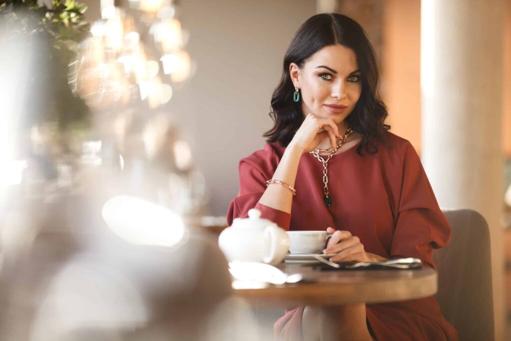A woman with dark hair sits at a table in a cafe, holding a cup. She wears a rust-colored top and jewelry, with a blurred foreground and warm lighting.